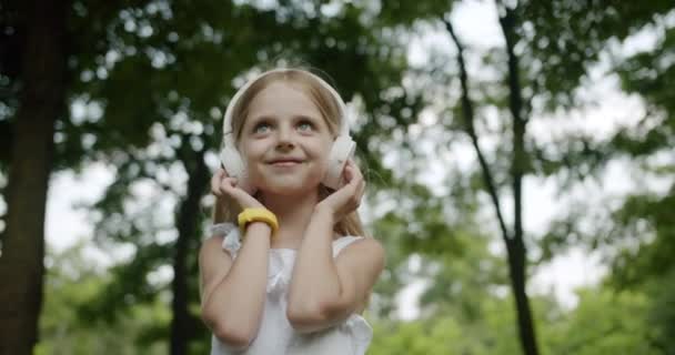 Happy child listens to music on modern white headphones in park and look up — Stock Video