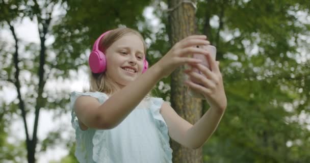 Tinédzser zenét hallgat rózsaszín fejhallgató a parkban, hogy online video hívás — Stock videók