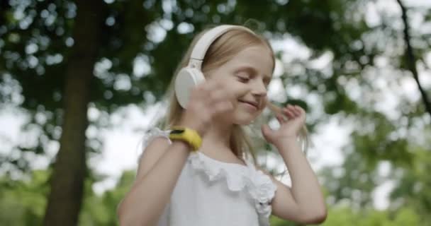 Criança feliz ouve música em fones de ouvido brancos modernos no parque e olha para cima — Vídeo de Stock