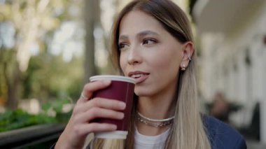 Genç bir kadın kahve fincanından yudumluyor, güneşli bir parkta huzurlu bir anın tadını çıkarıyor, yemyeşil ve yumuşak güneş ışığıyla çevrili. Sunny City Park Sahnesinde Kahve İçen Genç Kadın