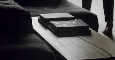 Modern bir Minimalist oturma odasında, siyah mobilyalar, ahşap masa ve kitapların olduğu yerleri kapatın. Adam masanın yanında yürür ve siluetler ve gölgeler yaratır..