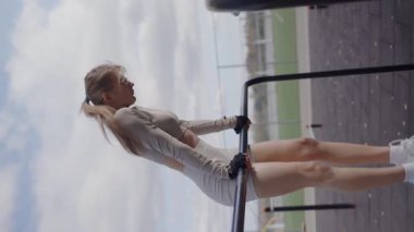 Genç aktif bir kadın açık hava spor salonunda paralel barlarda egzersiz yapıyor bulutlu bir gökyüzünün altında şık bir spor kıyafeti giyiyor. Güneşli bir günde dışarıda egzersiz yapan genç bir kadın. Dikey video.