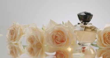 Siyah şapkalı zarif bir parfüm şişesi yansıtıcı yüzeyde güzel beyaz güllerle çevrilidir ve lüks ve romantik bir atmosfer yaratır. Taze Güllü Zarif Parfüm Şişe