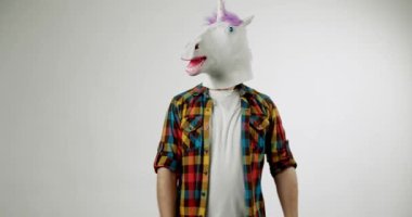 Tek boynuzlu at maskeli adam elleriyle bir şey gösteriyor. Komik tek boynuzlu at maskesi, ekose gömlek. Beyaz arka plan. Neşeli tek boynuzlu at. Tek boynuzlu at kostümü yap. Hayal gücü ve hayal gücü.