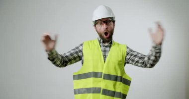 Şaşırmış bir inşaat işçisi heyecandan ellerini kaldırıyor. O, sert bir şapka, güvenlik gözlüğü ve yüksek görüş açısına sahip bir yelek giyiyor. Arkaplan koyu beyaz bir renktir.