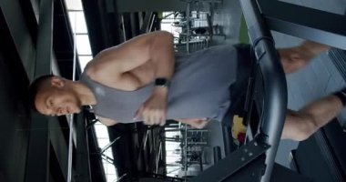 Genç bir adam, modern bir spor salonunda kürek çekerken yoğun bir şekilde odaklanır. Spor salonundaki Row Machine üzerinde çalışan odaklanmış adam. Dikey video.