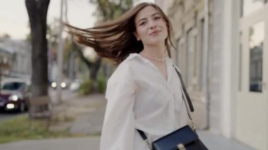 Beyaz bir bluz giyen, güneşli bir günde şehir caddesinde yürürken neşeyle gülümseyen genç bir kadının görüntüsü, ferahlatıcı bir şehir havası ekliyor. Caddede yürürken gülümseyen genç kadın.