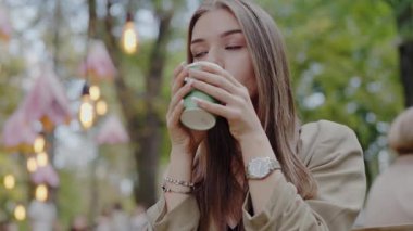 Genç bir kadın açık havada kahvesini yudumluyor. Etrafı yumuşak odaklı ışıklar ve yeşil ağaçlarla çevrili. Şehir parkında bir dinlenme anı yakalıyor. Genç Kadın Canlı Şehir Parkı 'nda Kahvenin tadını Çıkartıyor