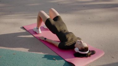Yoga minderinin üzerinde rahatlatıcı esnemenin tadını çıkaran genç bir kadın. Genç bir kadın, parkta yoga minderinde uzanırken kulaklıkları açık, gözleri kapalı güneşli bir günün tadını çıkarıyor..