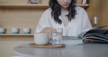 Beyaz bluzlu odaklanmış genç bir kadın modern ahşap bir masada dergi okurken suyunu yudumluyor. Arka planda şık mutfak eşyaları olan rahat bir ev ortamı.