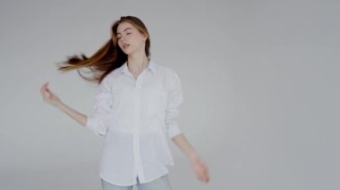 Uzun saçlı, nazikçe gülümseyen, gri arka planda günlük beyaz gömlek ve kot pantolon giyen genç bir kadının portresi. Günlük Beyaz Gömlek ve Kot Giymiş Gülümseyen Genç Kadın