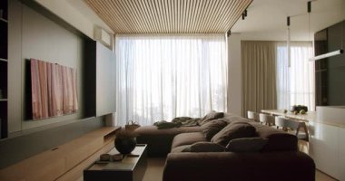 Modern oturma odasının içinde kahverengi kanepe, ahşap tavan ve katıksız perdeler var. Doğal ışık odayı dolduruyor, rahat bir atmosfer yaratıyor. Açık kat planı oturma odasını yemek alanına bağlıyor..