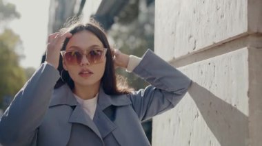 Güneş gözlüklü şık genç bir kadın işlek bir caddede güvenle yürüyor, saçlarına dokunuyor ve güneş ışığının tadını çıkarıyor. Güneş Gözlüklü Moda Genç Kadın Güneşli Şehir Gününün tadını çıkarıyor