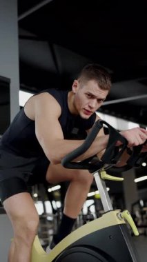 Ciddi bir ifadeyle odaklanmış genç bir adam modern bir spor salonunda sabit bir bisiklete biniyor. Azimli Genç Adam, Spor Bisikleti üzerinde Yoğun Egzersiz Yapıyor. Dikey video.