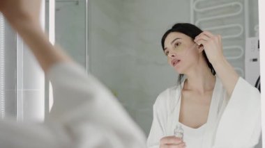 Beyaz cüppeli genç bir kadın modern bir banyoda aynanın önünde yüzüne cilt serumu sürüyor. Rahatlama ve kendine bakma rutini. Banyo Aynası Önünde Cilt Serumu Uygulayan Kadın