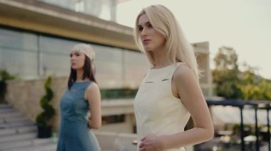 Modern bir binanın dışında poz veren zarif kadınlar. Biri beyaz elbiseli, diğeri mavi elbiseli ve başörtülü. Tarz, moda ve modern yaşam vurgulanıyor. İki Moda Kadın Pozu