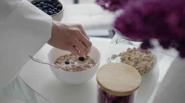 Bir kase granola ve süte taze yaban mersini ekleyen bir elin yakın çekimi. Sağlıklı kahvaltı kavramı ve besleyici bir yemek. Sağlıklı beslenme alışkanlıklarını geliştirmek için mükemmel..