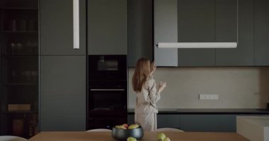 Pijamalı bir kadın modern mutfağından geçer ve bir dolap açar. Mutfak, koyu gri dolaplar ve açık ahşap bir masa ile minimalist bir tarzda dekore edilmiştir. Elinde bir bardak su tutuyor..