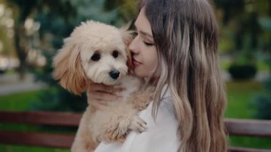 Genç bir kadın küçük tüylü köpeğini sevgiyle parkta tutuyor. Birlikte hassas bir anı paylaşıyorlar, sakin açık havada birbirlerine eşlik ederek eğleniyorlar..