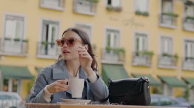 Güneş gözlüklü şık genç bir kadın açık bir kafede kahve yudumlarken arka plandaki şehir binalarında oturuyor. Şık Genç Kadın Şehir Kafesi 'nde Bir fincan kahvenin tadını çıkarıyor.