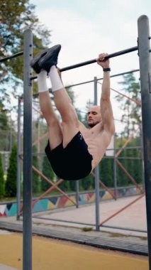 Kaslı bir adam parktaki barfiks çubuğunda sallanan bacağını kaldırır. Açık hava antrenmanı sırasında güç, zindelik ve çekirdek bağlılık gösteriyor..