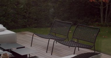 Arka bahçedeki ahşap bir güvertede oturan iki boş siyah hasır veranda sandalyesi. Yeşiller ve çalılar huzurlu bir atmosfer yaratıyor. Rahatlamak için mükemmel..