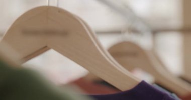 Dükkandaki ahşap askılara asılı kıyafetlerin yakın çekimi. Gömlekler yeşil, kırmızı ve mor. Dekor parlak ve havadar. Moda ve perakende için mükemmel..