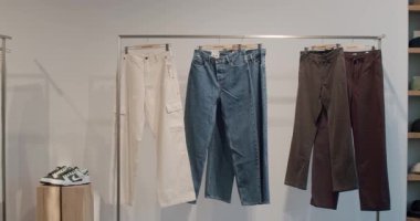 Bir giyim mağazasında çeşitli pantolon ve ayakkabılar sergileniyor. Beyaz kargo pantolonları, kot pantolonlar ve kahverengi pantolonlar bir askıda asılı, farklı stil ve renkler sergiliyorlar. Chisinau, Moldova - 18 Ocak 2025
