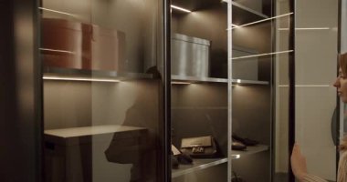 Genç bir kadın cam kapılı modern bir gardırop açıyor, içinde ayakkabılar, çantalar ve depolama kutuları olan organize raflar var. Gardırop dahili ışıklandırmayla aydınlatılmış.,