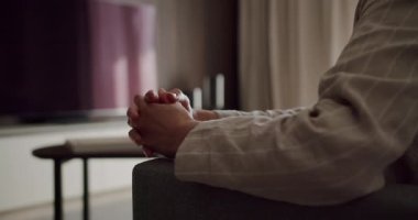Elleri sıcak bir oturma odasında sıkışık bir şekilde sandalyede oturan birinin yakın çekimi. Sahne bir rahatlama ve tefekkür hissi yayar. Sandalyeye oturmuş, elleri bağlı biri.