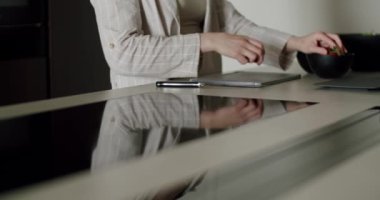 Çizgili ceketli bir iş kadını modern bir mutfakta dijital tablet üzerinde çalışırken sağlıklı bir atıştırmalık hazırlar. Çoklu görev anlayışı, sağlıklı yaşam tarzı ve teknoloji. Mutfak.