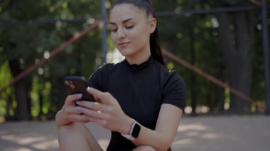 Atletik bir genç kadın spor kıyafetleri giyerek güneşli bir parkta antrenmana ara verirken akıllı telefonunu kontrol ediyor. Açık hava antremanı sırasında akıllı telefon kullanan atletik kadın