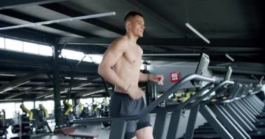 Modern bir spor salonunda koşu bandında koşan zinde bir genç. Genç kaslı bir adam iyi donanımlı bir spor salonundaki koşu bandında koşu yaparken kardiyo çalışmasına odaklanır..