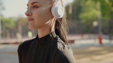Odaklanmış genç bir kadın, güneşli bir parkta koyu renk bir ceket giymiş beyaz kulaklıkla müzik dinliyor. Huzur dolu bir an. Güneşli bir günde Parkta Müzikten Hoşlanan Genç Kadın