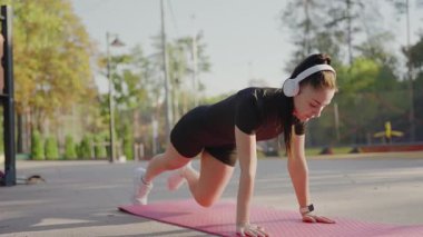 Kulaklıklı, dinamik yoga pozisyonunda, açık havada pembe bir minder üzerinde duran odaklanmış genç bir kadın spor ve dengeyi gösteriyor. Dışarıda Dinamik Yoga Pozu Veren Genç Kadın