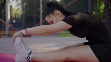 Atletik giysiler içindeki odaklanmış genç bir kadın bir parktaki yoga minderine dizlerini uzatarak sağlıklı bir yaşam tarzı ve formda kalma alışkanlığı sergiliyor. Açık hava parkında Hamstring Esnemesi Yapan Genç Kadın