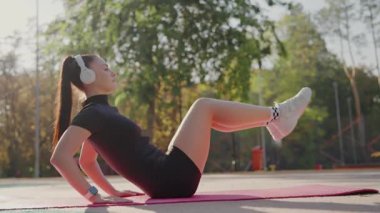 Beyaz kulaklıkla müzik dinlerken, parkta pembe bir minder üzerinde egzersiz yapan zinde bir genç kadın. Açık havada egzersiz yapan, güneşli bir günde müzikten zevk alan genç bir kadın.