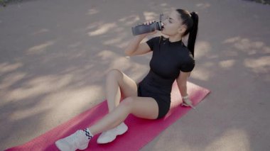 Sporcu formalı genç bir kadın yoga minderinde oturup antrenmandan sonra su içiyor. Sağlıklı yaşam tarzı ve spor konsepti. Yoga Mat 'te Dinlenen ve İçen Genç Kadın
