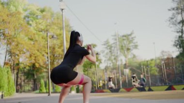 Spor kıyafetleri içinde odaklanmış genç bir kadın parkın dışında, kulaklık takıyor ve güneşli bir günün tadını çıkarıyor. Aktif Kadın Kulaklıkla Parkta Esneme Yapıyor