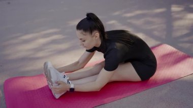 Odaklanmış genç bir kadın, barışçıl bir açık hava parkında pembe bir yoga minderi üzerinde esneme egzersizleri yapıyor. Spor giysisi ve fitness saati takıyor. Açık havada Pembe bir mindere uzanan genç kadın.