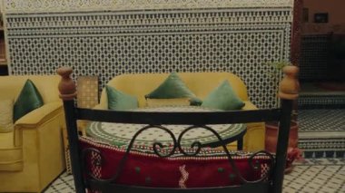 Sarı kanepe, yeşil yastıklar, mozaik masa ve desenli duvar kiremitleri olan sakin Fas avlusu sahnesi. Bu huzur, rahatlama ve egzotik seyahat hissi uyandırıyor..