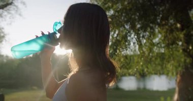 Spor yapmaya ara veren, elinde şişe tutan ve güneşli bir günde parkta su içen atletik kadınların yakın çekimi. Genç bayan dışarıda idmandan sonra ferahlatıcı. Temiz maden suyu.