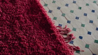 Canlı, peluş, koyu kırmızı Fas halısının püsküllü yakın çekimi. Kısmen karmaşık, geleneksel Fas fayanslı zemini beyaz, turkuaz ve zümrüt yeşiliyle kaplıyor. Geometrik desenler