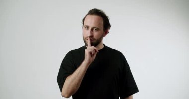 Dudakta Parmaklı Adam Sessiz İşaret Gösteriyor. Siyah tişörtlü Sakallı Adam Beyaz Arkaplanda Sessizlik Hareketi Yapıyor. Sır, Komplo, Gizlilik Konsepti. Vücut Dili, Sözsüz İletişim. 4K.