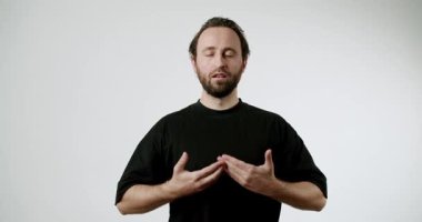 Siyah tişörtlü bir adam beyaz arka planda duruyor, gözleri kapalı, ellerini yavaşça hareket ettiriyor. Farkındalık ve meditasyon yapıyor, huzur dolu bir atmosfer yaratıyor.