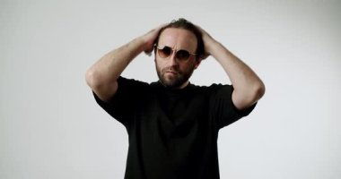 Sakallı ve güneş gözlüklü bir adam saçını beyaz bir arka plana göre ayarlar. Siyah bir tişört giyer ve doğrudan kameraya bakar. Kendine güveni ve tarzı aktarır..