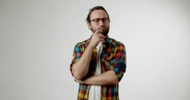 Gözlüklü ve sakallı düşünceli bir adam çenesini okşuyor. Beyaz bir arka fona karşı renkli bir ekose gömlek giymiş. Bu video derin düşüncelere sahip.,