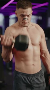 Kas yığını bir adam, spor salonunda mor neon ışıkları olan dambılları kaldırır. Üstsüz ve antrenmanına odaklanmış durumda. Dambılları gayretle kaldırır, pazılarını çalıştırır ve kas geliştirir..