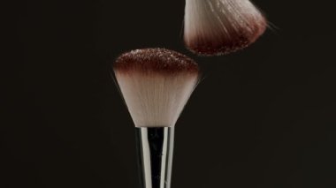 Havanın ortasında çarpışan iki kozmetik fırçanın yavaş çekim klipsi koyu bir arkaplana karşı yüz pudrası bulutu yaratıyor, makyaj uygulamasını ve güzellik kavramlarını sembolize ediyor.