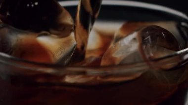 Koyu bir zemin üzerinde buzla dolu bir bardağa dökülen siyah kahvenin yakın çekimi, serinletici bir soğuk içeceği yavaş çekim ile sıcak bir yaz günü için mükemmel kılıyor.